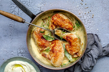 Braised pieces of chicken with dijon mustard sauce in a copper pan with herb garnish on light, grey background, landscape style, overhead shot with grey tea towel and a side of mash