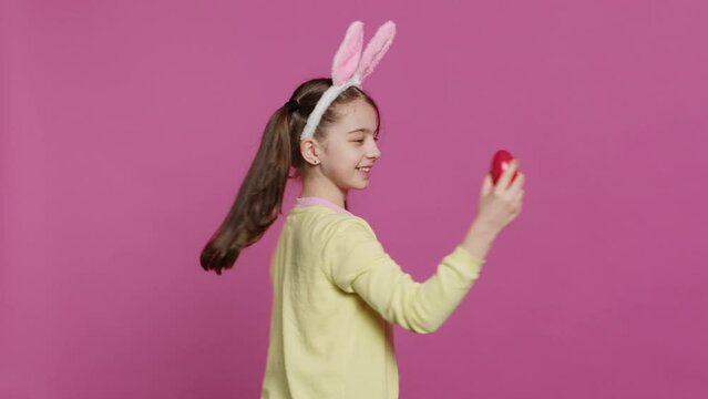 Enthusiastic small child spinning around in front of camera, presenting her red painted egg for easter sunday festivity. Cheery toddler with bunny ears twirling and doing a pirouette. Camera A.