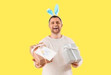 Happy young man in bunny ears with gift boxes and greeting card on yellow background. Easter celebration
