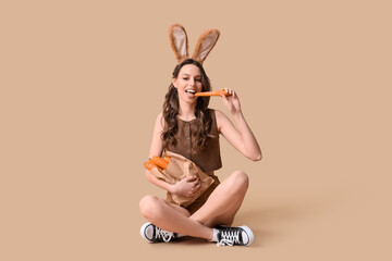 Happy young woman in Easter bunny ears headband with paper shopping bag eating carrot on beige...