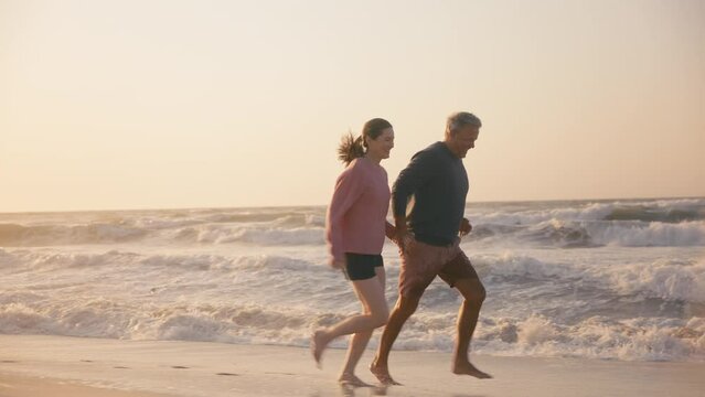 Camera tracks loving retired senior couple on vacation running along beach shoreline splashing through waves and holding hands at sunrise - shot in slow motion