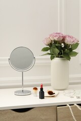Mirror, cosmetic products, jewelry and vase with pink roses on white dressing table in makeup room