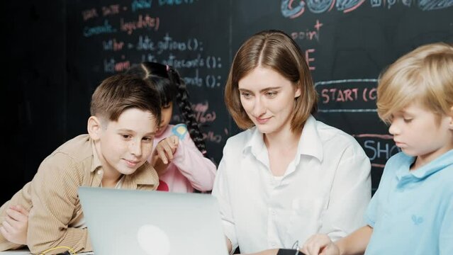 Caucasian teacher using laptop and explain about engineering system while diverse student holding electronic equipment in front of blackboard with prompt or code written in STEM class. Erudition.