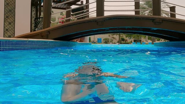 Lockdown shot of happy woman emerges from water in swimming pool. Female enjoys clear water at resort pool. She is spending leisure time during holiday.
