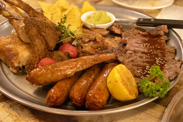 Close-up of barbecue platter under warm light, including sausages, steak, chicken, lamb chops, etc.