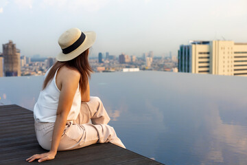 Pretty tourist woman is relaxing and enjoying her rooftop swimming pool view with cityscape...