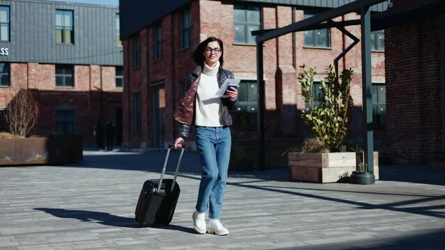 Full length of adult female pulling baggage on sidewalk and heading to departure station with pack of documents. Caucasian woman leaving rent apartment after journey on street with brick buildings.