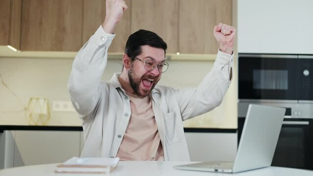 Surprised caucasian man looking at screen of wireless laptop and making winner gesture. Cheerful bearded male expressing positive emotions from online recieved news. Concept of win and triumph.