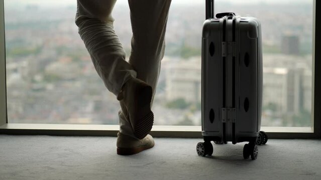 Man stands by window, suitcase beside him. We see only his legs in suede shoes and wheeled bag. Man raises his left leg, scratching calf of his right foot. He's bit nervous about upcoming trip