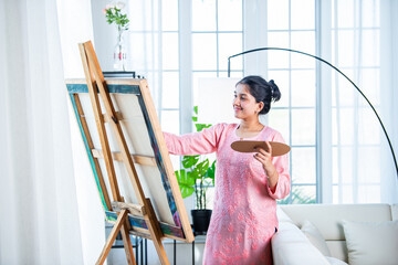 Cheerful Indian young artist girl painting picture at easel in living room