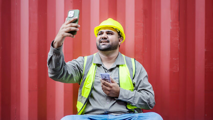 Young asian container male worker or dock foreman sit and rest near the container. I'm on a video call by phone with my family, happy that I received my salary today, earning or profit