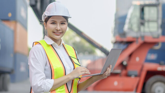 Asian engineer woman working in container port terminal. Young businesswoman process order and product at warehouse logistic in cargo freight ship for import and export, look at camera and smile