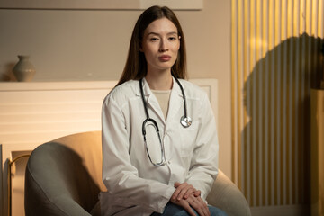 A young medical woman in a white lab coat and with a stethoscope sits in a well-lit modern office,...