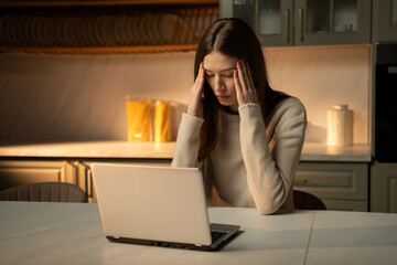 A young woman is sitting at the kitchen table, clearly upset, with tears in her eyes, as she looks up from her work at her laptop for a moment