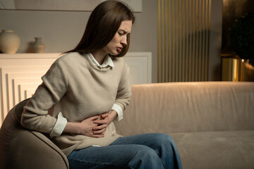 A woman is seated on a sofa clutching her abdomen with a pained expression, suggesting she is...