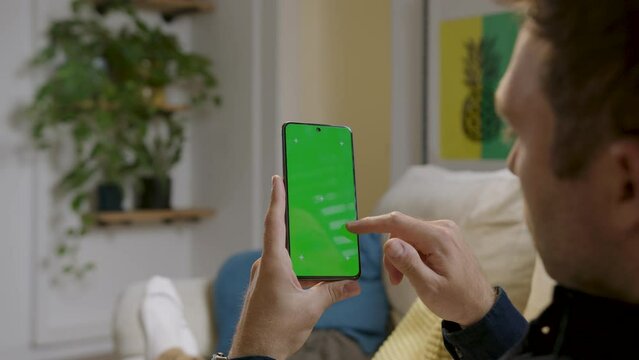 Man comfortably seated at home, holding a smartphone with green screen and interacting with it as if he were using it