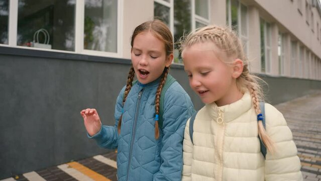 Two little European girls sisters best friends classmates walking street city outside talking chatting discussion schoolgirls school lessons gossip communication education relations vacations sibling