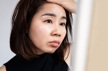 worried Asian woman have problem with oily skin on t zone of her face looking in a mirror
