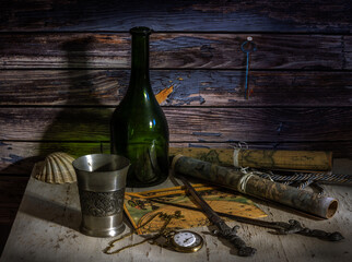 Antique nautical maps on the table pressed with daggers and seashells. Empty glass bottle and metal glass. Pocket watch with chain. Marine theme.