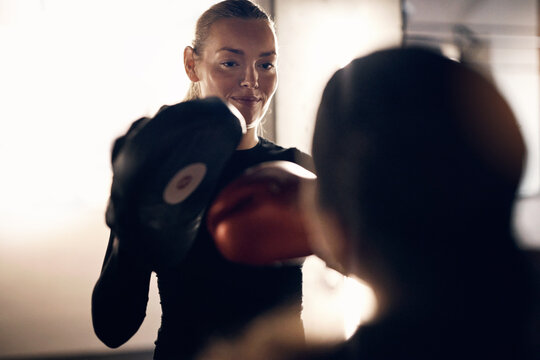 Over the shoulder view of a fit young woman holding hands pads for a female friend practicing her punching during a boxing workout in a gym