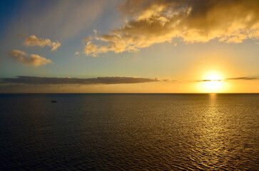 Golden sunset at sea, sailing boat on horizon, reflections of sunrays, sunset sky with clouds