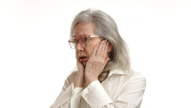 Captured in stark detail, an elderly woman's face is frozen in fear, clutching her head against a plain white backdrop, depicting pure terror and anxiety. Camera 8K RAW. 