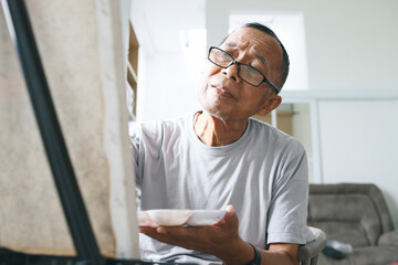 Senior Asian man wear glasses, sit and painting picture inside of the house. Happy retirement lifestyle concept.