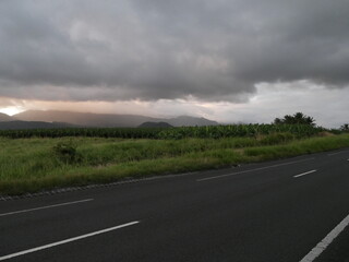 Naklejka premium empty highway and tropical field in basse terre, guadeloupe. Asphalt road with empty lanes and mountain background with storm clouds in dramatic sky