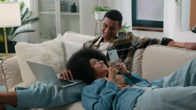 African American couple man with laptop computer sitting on couch home woman chatting smartphone browsing mobile phone laying on male knees girl guy gadget addict internet addiction smiling family