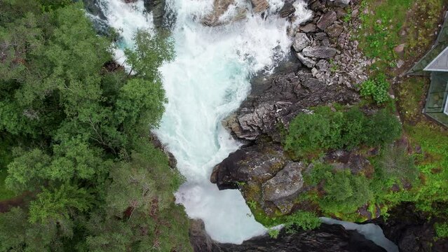 Top down shot, pushing in on a raging waterfall in Norway. White water rapids crash forcefully against a narrowing in the rocks before gushing down a cliff. Lush trees and grass surround the waterfall