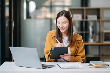 Young beautiful woman typing on tablet and laptop while sitting at the working white table in office