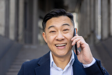 Smiling asian businessman in suit engaging in a pleasant phone conversation outside