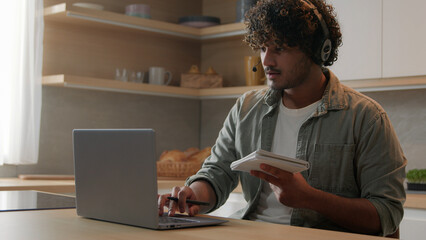 Indian man in headset look app pc browsing typing laptop write ideas questions notes review...