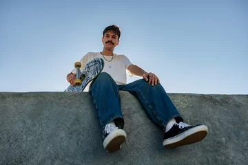 Tapeten Low angle of young male skater in casual outfit sitting on ramp with skateboard against blue sky © Kostiantyn