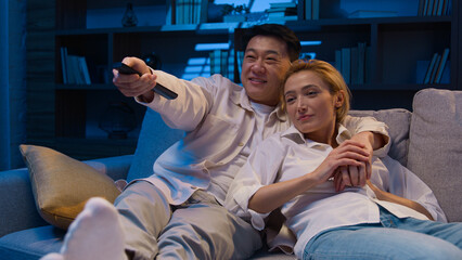 Multiracial couple watch movie at night evening diverse married Asian man and Caucasian woman happy family multiethnic Korean husband and wife watching TV television relaxing at home couch cuddling