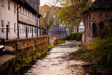 A serene creek flows through an old town, its waters glistening at dusk, flanked by traditional half-timbered houses and a cobblestone path