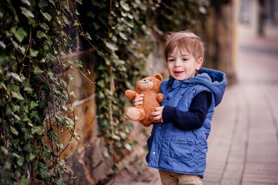 A smiling child holds his teddy bear close, a picture of happiness against a natural backdrop of climbing ivy.