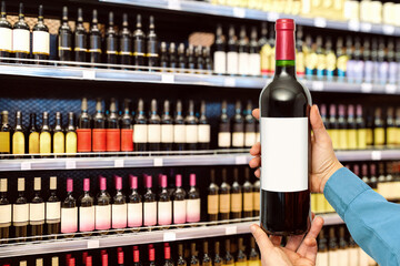 Wine bottle with empty label in man's hands in front of wine bottles on shelves on liquor store. 