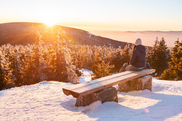 Sudetes, a young woman sits on a bench overlooking the mountain landscape at sunset, view from a winter hiking trail.