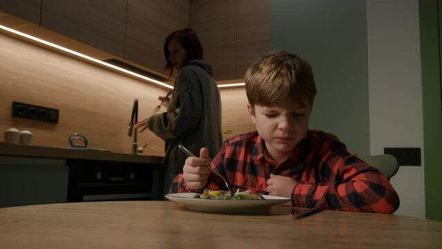 A picky eater protests against a bland meal while his mom prepares food in the kitchen, capturing a common family struggle