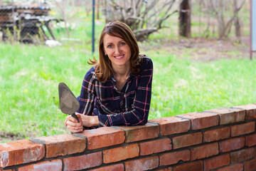 a young female bricklayer with a trowel in her hands is smiling, leaning against a brick wall