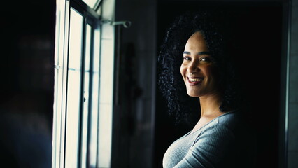 Fototapeta premium Happy Portrait of a young black Brazilian woman turning head to camera smiling standing by window at home. Curly hair South American adult girl smiling