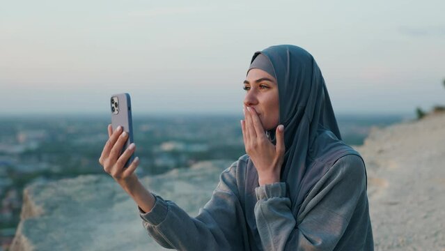 An attractive woman in a hijab is practicing yoga on the slope of a mountain. The girl is communicating via video call on her phone and sends an air kiss to the person she's talking to.