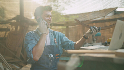 Young carpenter talking on smartphone and using laptop while working on woodworking in the furniture factory. Carpenter and woodcraft concept