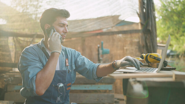Young carpenter talking on smartphone and using laptop while working on woodworking in the furniture factory. Carpenter and woodcraft concept