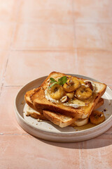 Sweet toast with cream cheese, caramelized banana, nuts and honey on beige tile background, hard morning light. Text space