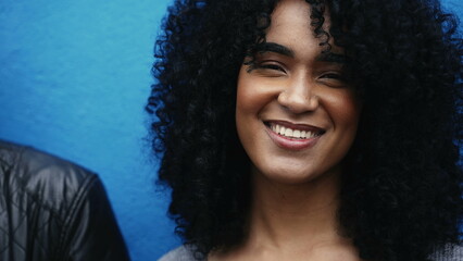 One joyful young latina black woman with curly hair smiling at camera on blue backdrop in city environment. close-up face of South American person of African descent - obrazy, fototapety, plakaty