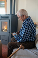 Old man in his eighties looking at mobile phone screen, sitting on sofa next to pellet cooker....