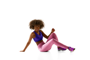 Joyful woman in purple gym wear resting, sitting on floor, looking at camera with bright smile against white studio background. Concept of sport, mourning routine, active and healthy lifestyle, energy