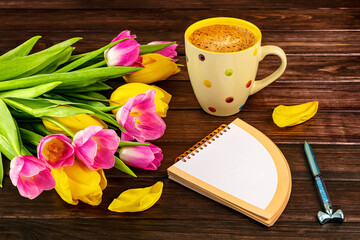 Spring bouquet of yellow and pink tulips, white polka dot cup of coffee, colorful notebook, pen on dark wooden background. Spring still life